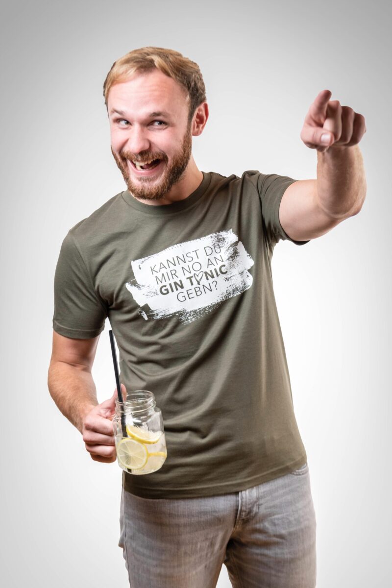 Mann mit T-Shirt und einem Gin-Tonic in der Hand. Auf dem Shirt steht "Kannst du mir bitte no an Gin Tonic gebn!"